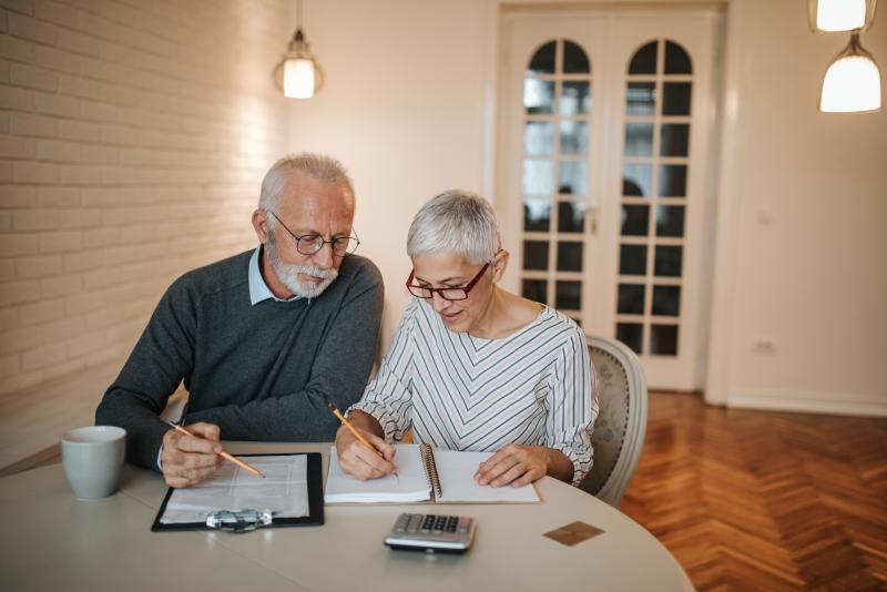 An elderly couple sits at a table in front of a computer and a calculator.