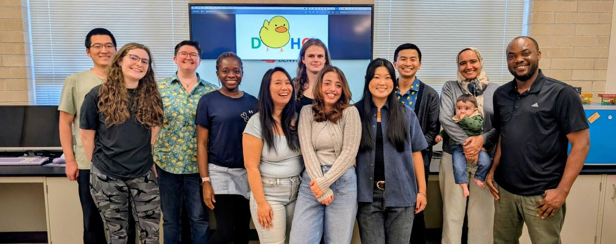 12 members of the University of Denver Human-Computer Interaction Group standing together smiling inside a room. Behind them is a pixel art logo which reads "DUHCI" with a duck in place of the letter "U"